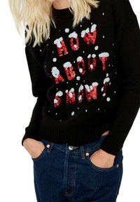 Musta neulepaita, jossa on punaisista paljeteista teksti "MITÄ SANOTTAISIIN LUMESTA?" ja valkoisia tekstuurisia yksityiskohtia, jotka muistuttavat lumihiutaleita. Yhdistettynä sinisiin farkkuihin.