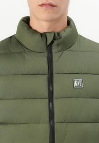 Zelena zimska jakna s visokim ovratnikom, zatvaranjem na patentni zatvarač i šavovima. Ima crni patentni zatvarač i GAP logotip na prsima.