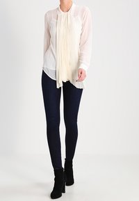 Žena v průhledné bílé blůze s dlouhými rukávy a plisovanými detaily v přední části, tmavých skinny džínách a černých kožených kotníkových botách na podpatku.