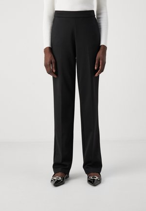 Black, high-waisted trousers with a straight cut, featuring a smooth texture. Worn with pointed, patent black shoes adorned with metallic accents.