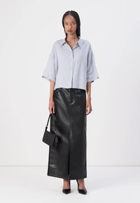 Camicia di lino azzurro chiaro con maniche corte, abbinata a una lunga gonna di pelle nera. Tiene una piccola borsa nera, accentuata da tacchi neri.