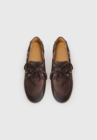 Sapatos de barco em pele castanha com biqueira arredondada, estilo de atar e costura contrastante. Textura suave com ilhós de metal e laços em pele.