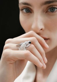 Anillo de plata diseñado como un cocodrilo con escamas texturizadas y una piedra negra como ojo, usado en un dedo contra un fondo neutro.