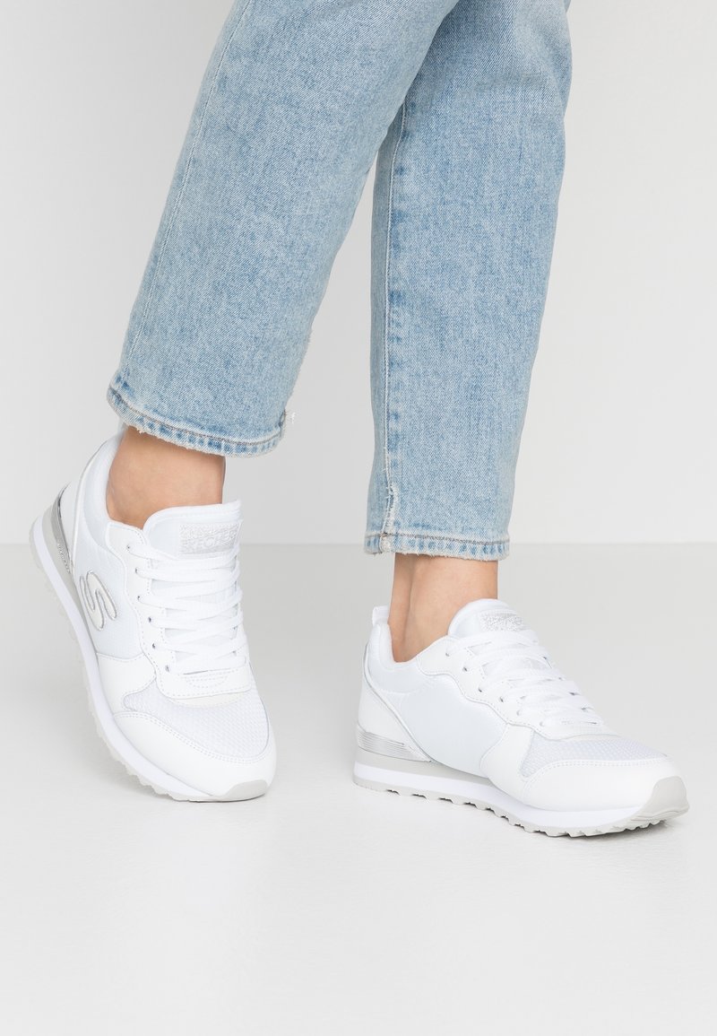 Personne portant un jean bleu clair et des baskets blanches debout sur un sol gris clair sur un fond blanc uni.
