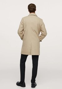 Trench beige avec une fente au dos, des épaulettes et des boutons noirs, porté avec un pantalon noir et des chaussures noires épaisses.