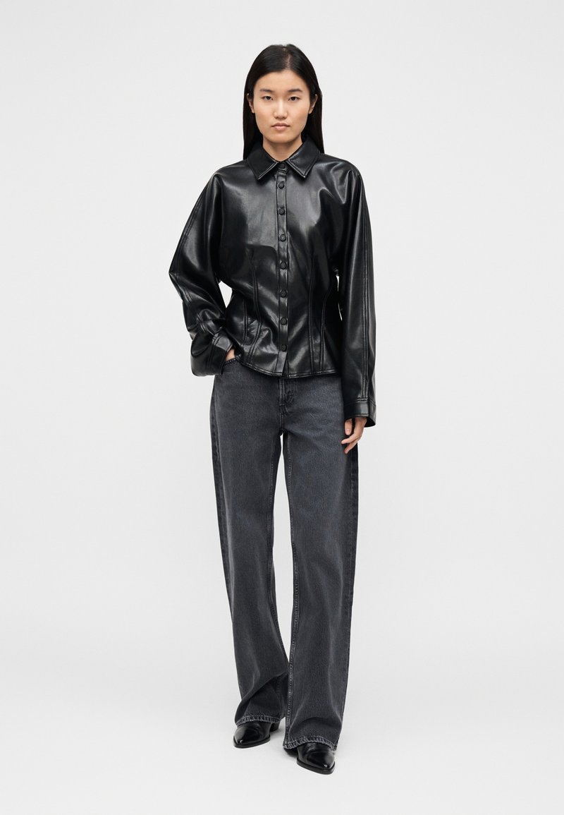 Chemise en cuir noir avec fermeture à boutons, col pointu et manches longues. Associée à un jean gris foncé à larges jambes et des bottines noires.