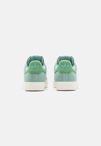 Green suede sneakers with a smooth texture, featuring a white rubber sole and embossed logo on the heel. Clean, minimalist design.