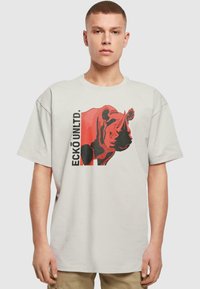 Lichtgrijze t-shirt met een groot rood en zwart neushoornontwerp en "ECKŌ UNLTD." tekst aan de linkerkant. Korte mouwen en een ontspannen pasvorm.