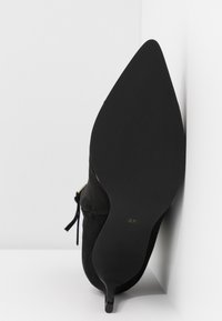 Chaussure à talon stiletto en daim noir à bout pointu, inclinée contre un coin blanc, taille 37 marquée sur la semelle.