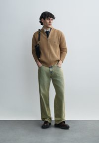 Μπεζ ριμπ μπλουζάκι με φερμουάρ, ανοιχτοπράσινο παντελόνι κοτλέ με φαρδιά μπατζάκια και σκούρα παπούτσια. Μαύρη τσάντα ώμου με λουρί. Ουδέτερο φόντο.