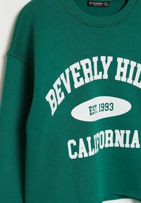 Felpa in pile verde con testo bianco che recita "BEVERLY HILLS CALIFORNIA EST. 1993." Collo a giro con dettagli a coste.