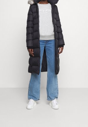 Zwarte pufferjas met een capuchon en nep-bontafwerking, gecombineerd met een lichtgrijs sweatshirt en brede blauwe jeans, afgemaakt met witte sneakers.