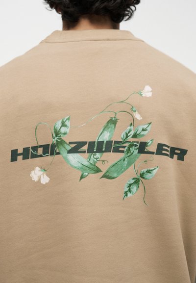 Άτομο που φοράει μπεζ πουκάμισο με σκούρο πράσινο κείμενο και γραφικά με λοβούς μπιζελιού και λευκά λουλούδια στην πλάτη.