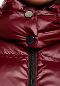 Bordo ocieplana kurtka o błyszczącej fakturze, z wysokim kołnierzem, czarnym zamkiem błyskawicznym i dwoma metalowymi zatrzaskami przy szyi.