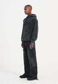 Sudadera negra de gran tamaño con un diseño texturizado, combinada con pantalones cargo negros holgados que cuentan con bolsillos cargo y acentos bordados. Zapatillas negras.