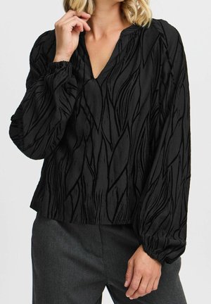 Vrouw draagt een zwarte blouse met lange mouwen met een subtiel bladvormig patroon en grijze broek, hand bij haar gezicht, tegen een effen achtergrond.