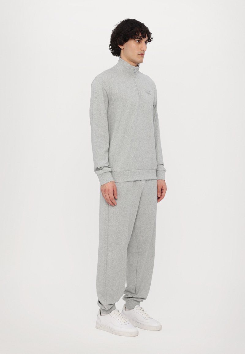 Sweatshirt zippé gris et pantalon de survêtement assorti en tissu doux, avec une coupe décontractée et des poignets élastiques, associé à des baskets blanches.