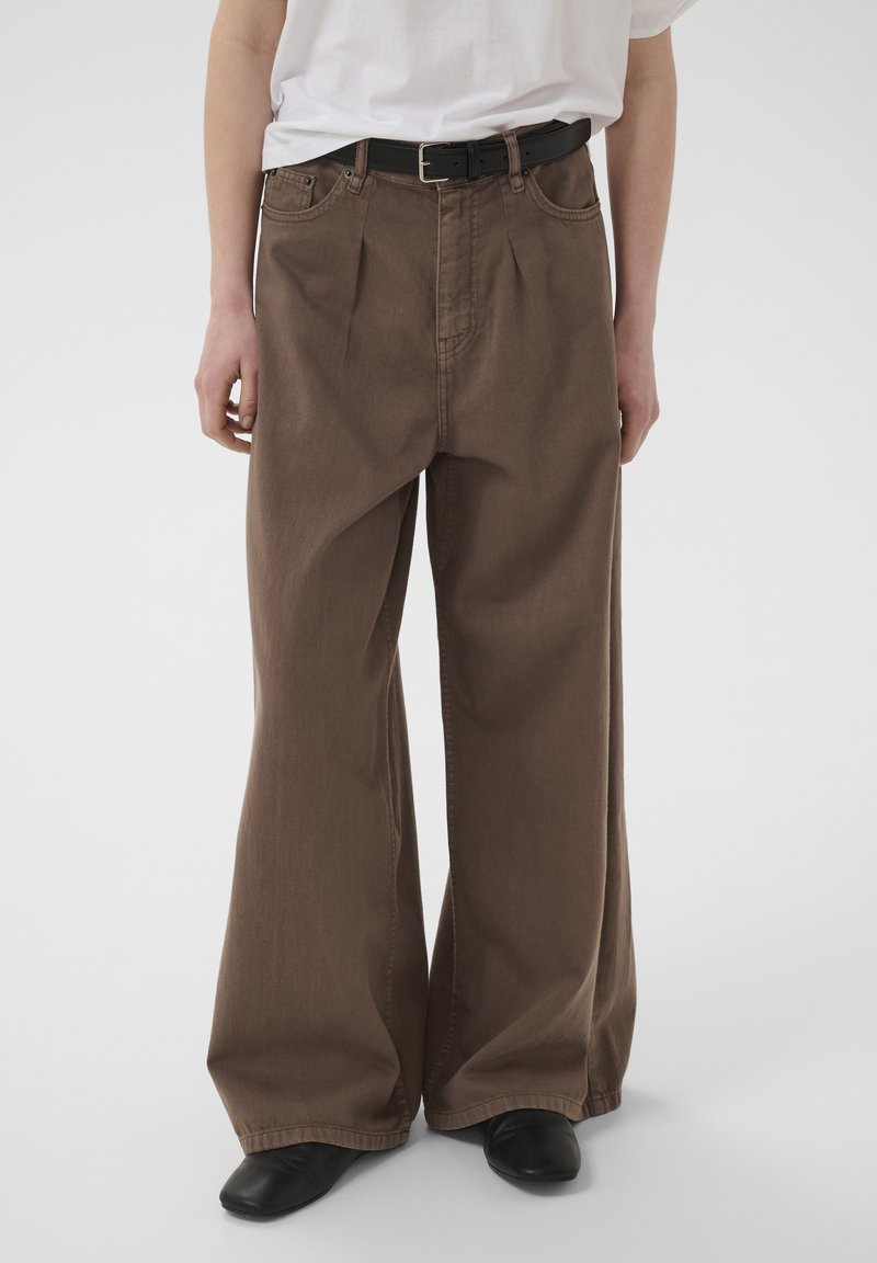 Personne portant un pantalon marron à jambes larges, une ceinture noire, une chemise blanche et des chaussures noires, debout devant un fond clair uni.