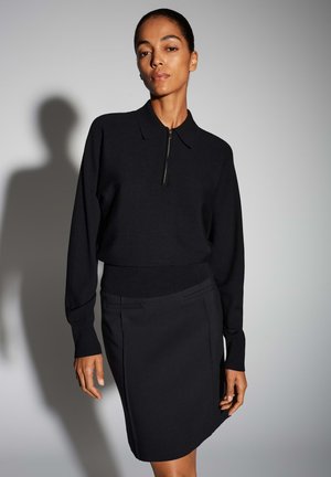 Schwarzes Strickoberteil mit Kragen und Reißverschluss, kombiniert mit einem figurbetonten schwarzen Rock mit Seitentaschen und klaren Linien. Glatte Textur.