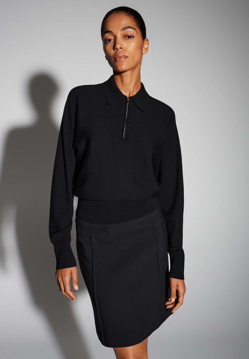 Haut en maille noire avec col et fermeture éclair, associé à une jupe noire ajustée avec poches latérales et lignes épurées. Texture lisse.