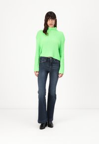 Pulover ribbed neon verde cu un guler înalt, asociat cu blugi evazați de culoare albastru închis și cizme negre până la gleznă. Culori solide, design ajustat.