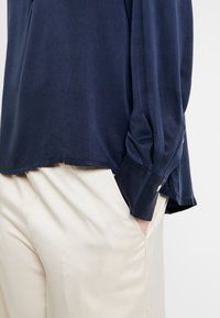 Top navy blu, a maniche lunghe, in tessuto morbido con una vestibilità rilassata e dettagli alle maniche con bottoni, abbinato a pantaloni chiari in tessuto liscio.