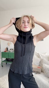 Haut sans manches noir à côtes avec un col haut et des cordons de serrage, tissu gris texturé, forme ajustée, porté avec un pantalon noir.