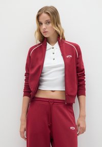 Giacca sportiva rossa con zip e piping bianco, dotata di un colletto alto. Abbinate a pantaloni jogger coordinati, che mostrano un design elegante e una texture morbida.