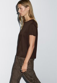 Brun kortærmet t-shirt med en afslappet pasform og rund halsudskæring, parret med afdæmpede brune ruskindsbukser. Glat tekstur og enkelt design.