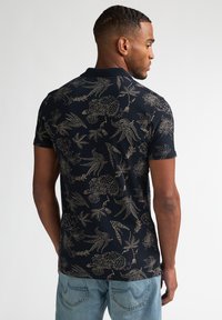 Marineblå poloshirt med gyldent blomster- og fuglemønster, korte ærmer og klassisk krave. Bærer lyse denimjeans.