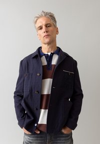 Navy textured jacket with button closure, featuring dual front pockets and a small chest pocket, worn over a striped polo in red, white, and blue.