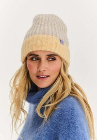 Dzianinowa czapka beanie w jasnoszarym i żółtym kolorze z prążkowanym wzorem, z podwiniętym mankietem i małą fioletową metką. Materiał z mieszanki wełny o fakturze.