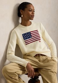 Femeie cu păr închis la culoare prins în coc, purtând un pulover crem cu un model tricotat cu steagul american și pantaloni bej, așezată pe un taburet de lemn.