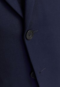 Námornícky modrý blejzer s hladkým povrchom, ktorý má klopový golier a čierny gombík. Záber zblízka ukazuje šitie a detail gombíka.