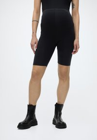 Persoană purtând pantaloni scurți negri de biker cu talie înaltă și ghete negre robuste, în fața unui fundal simplu și deschis la culoare.