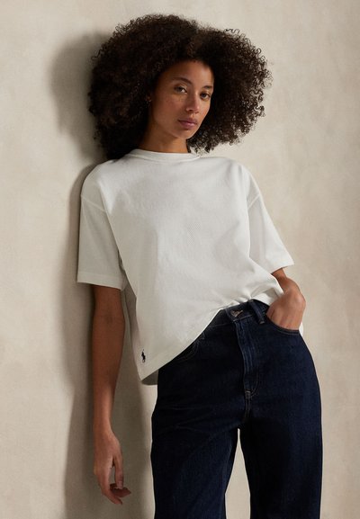 Kobieta z kręconymi włosami, ubrana w luźną białą koszulę z krótkim rękawem i ciemnoniebieskie dżinsy, opierająca się o beżową ścianę, ręka w kieszeni.