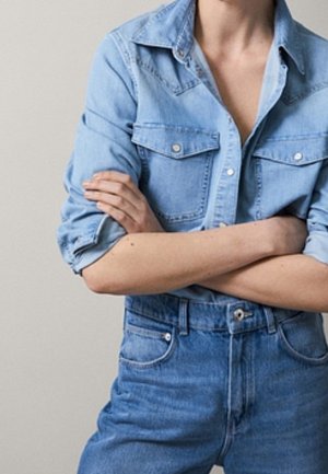Person wearing light blue denim shirt with sleeves rolled up and high-waisted blue jeans, arms crossed at chest.