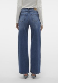 Weite blaue Jeans aus Denim mit hoher Taille, ausgestattet mit Gesäßtaschen und einem Leder-Logopatch an der Taille. Glatte Textur, ohne Verzierungen.