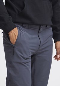 Grå bukser laget av myk stoff med slim fit. Har glidelås-lommer og en jevn tekstur, kombinert med en svart hettegenser.