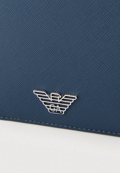 Tengerészkék texturált anyag ezüst emblémával, amely egy stilizált sast és középen a "GA" betűket ábrázolja. Hozzáillő varrással díszítve.