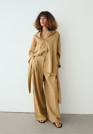 Kvinde står mod en ensfarvet væg iført løs beige skjorte med pinstriber og vidde bukser, barfodet i sorte sandaler, hænderne i lommerne.