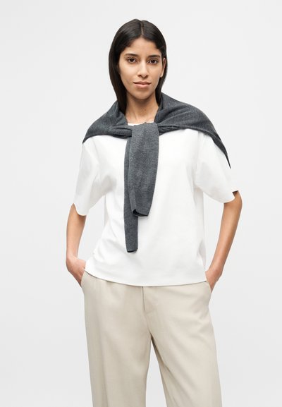 Λευκή μπλούζα με κοντά μανίκια συνδυασμένη με πλατύτερο παντελόνι σε ανοιχτό μπεζ χρώμα. Ένα σκούρο γκρι πλεκτό πουλόβερ είναι δεμένο γύρω από τους ώμους.