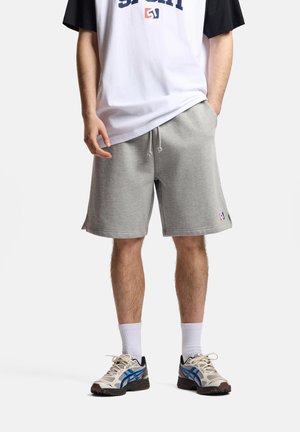 Mężczyzna noszący szare szorty na sznurku, białą i czarną koszulkę, białe skarpetki i beżowo-niebieskie buty do biegania, stojący z jedną ręką w kieszeni.