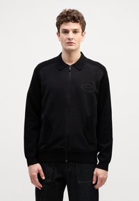 Črn zip-up jopič s ovratnikom, ki ima dva stranska žepa in teksturirano tkanino. Logotip na levi prsni strani, priložnostna prileganje.