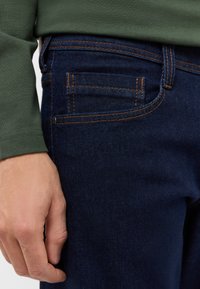 Ciemnoniebieskie jeansy z denimu z pomarańczowymi szwami, z przednią kieszenią i zbliżeniem na rękę spoczywającą na obszarze uda.