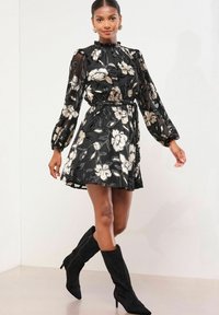 Rochie florală neagră cu mâneci lungi transparente, talie strânsă, guler înalt și accente florale aurii. Purtați cu cizme negre din suede, până la genunchi.