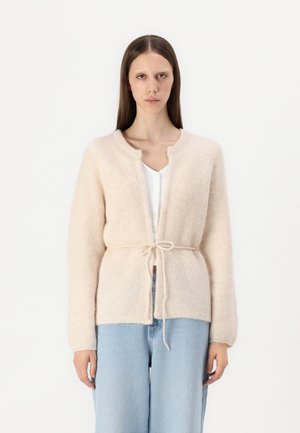 Blød beige cardigan lavet af tekstureret strikmateriale, med en frontknaplukning, åben design og lange ærmer, parret med lyseblå jeans.