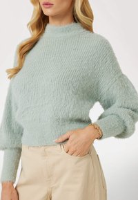 Šviesiai žalia megzta megztinis su aukštu kaklu ir laisvu siluetu. Yra faktūruotas audinys ir raukšlėti rankogaliai, derinamas su smėlio spalvos kelnėmis.