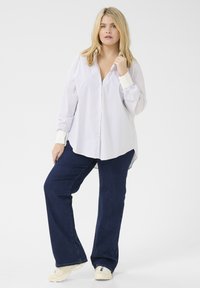 Camisa de botones blanca y azul a rayas con mangas largas y dobladillo curvado, combinada con jeans acampanados de azul oscuro y zapatillas blancas.