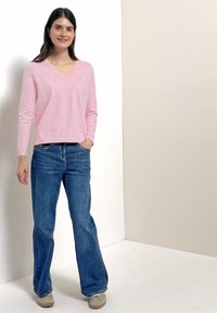 Femme debout, portant un pull rose clair à manches longues en V, un jean bleu et des baskets beiges, souriante devant un fond uni.